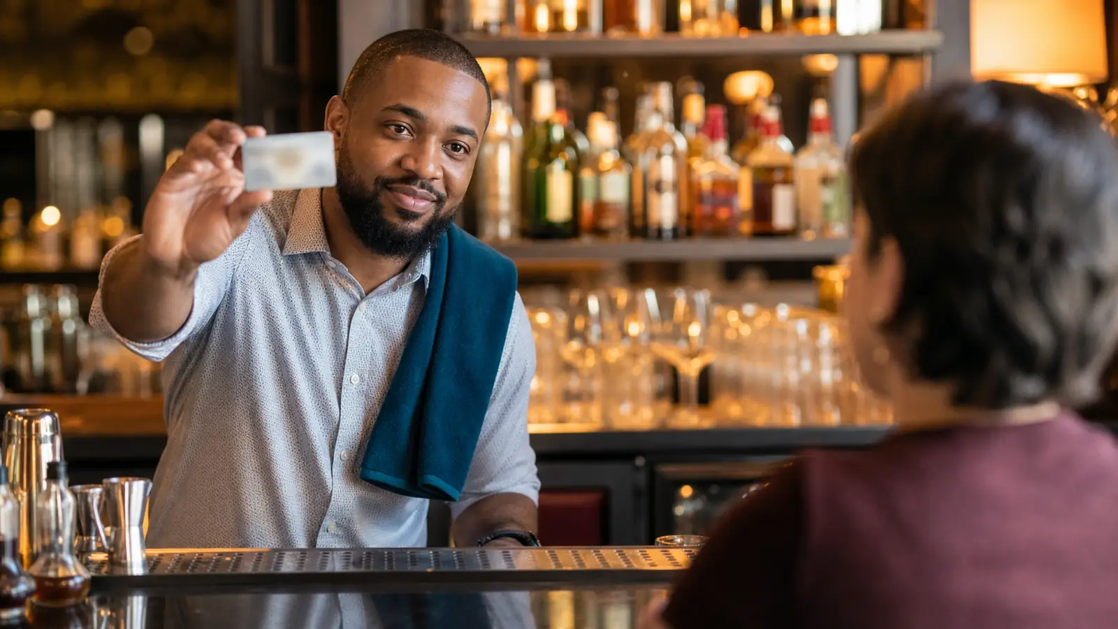 Bartender with a teal towel on his shoulder politely checking an unreadable ID card at a warm bar