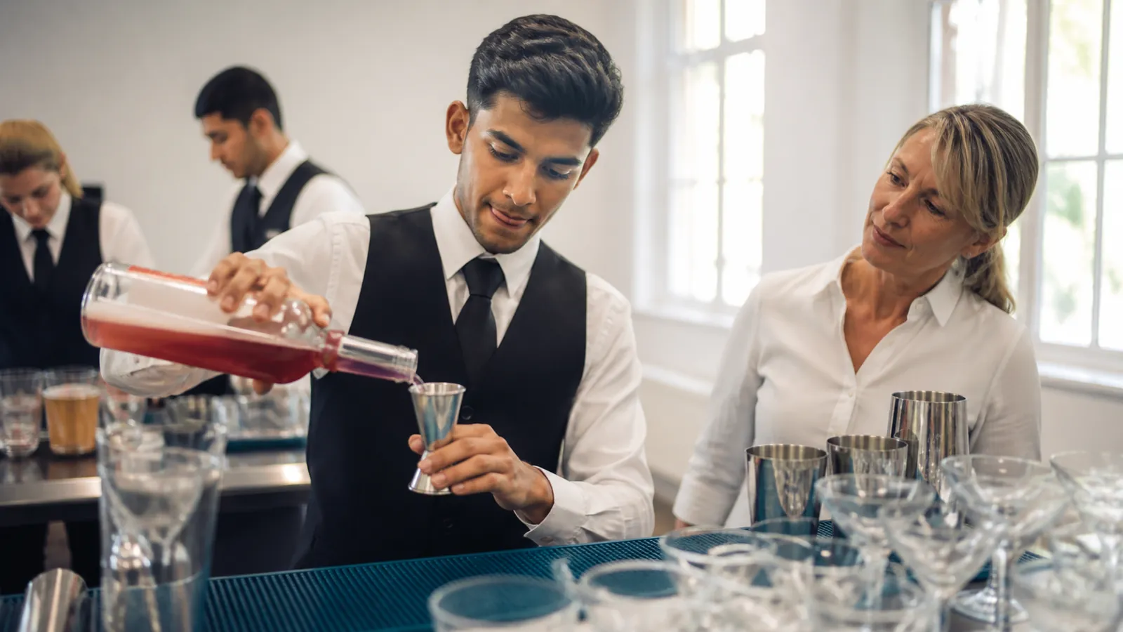Trainee free-pours practice liquid into a jigger over a teal bar mat as a trainer watches in a bright classroom