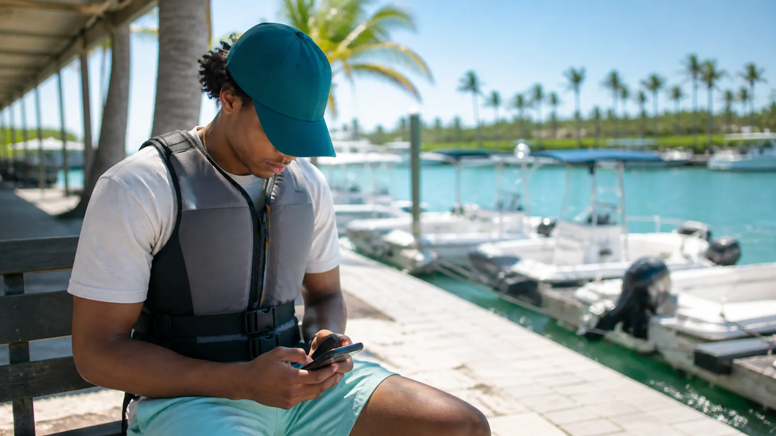 Young man in a life jacket studies a glowing phone on a sunny Florida dock, teal cap shading his eyes