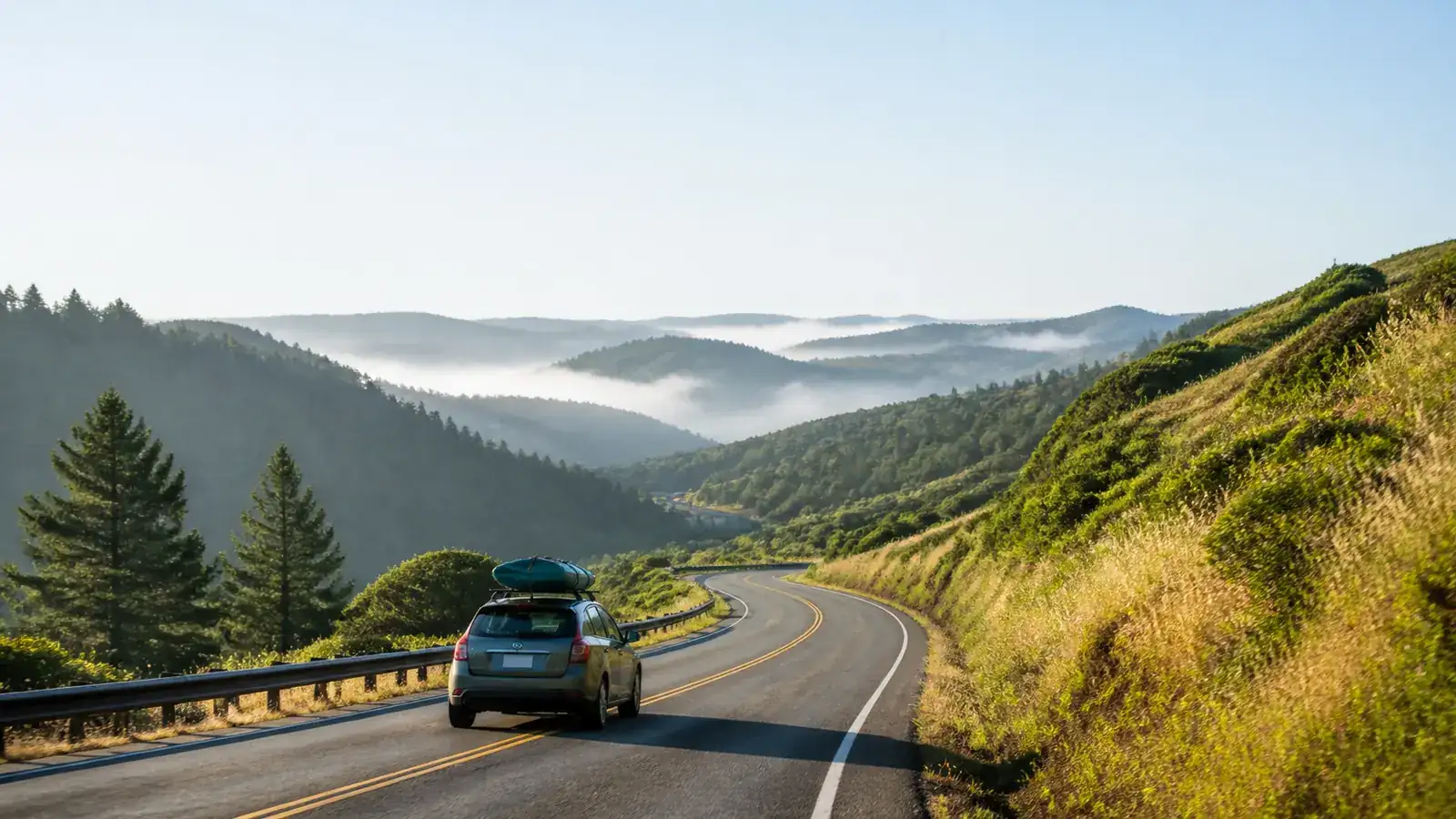 Small car with a teal kayak on its roof driving away down a curving two-lane road through green hills
